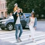 A young family crossing a city street at a crosswalk. The father carries a toddler on his shoulders as the mother walks beside them, all smiling and enjoying a sunny day out.