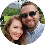 A smiling couple poses outdoors, with the man wearing sunglasses and a light blue shirt, and the woman with long, wavy hair. Lush greenery and mountains form the backdrop.