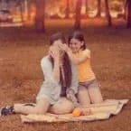 Mother and daughter playing peek-a-boo on a picnic blanket in a sun-dappled park. The girl covers her mom's eyes, both smiling.