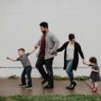 Family of four walks happily along a sidewalk, holding hands. The parents wear casual clothes and cardigans, while the children wear a t-shirt and dress. Light reflects off a white wall in the background.