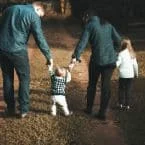 A family walks together at dusk, holding hands. A small child takes tentative steps between their parents, while an older child walks alongside the mother.