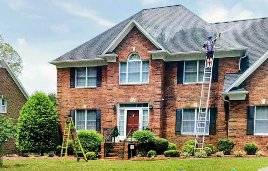 Professional house washing in progress: A worker on a ladder power washes a large brick house with dark shutters, while a smaller ladder stands on the lawn below.