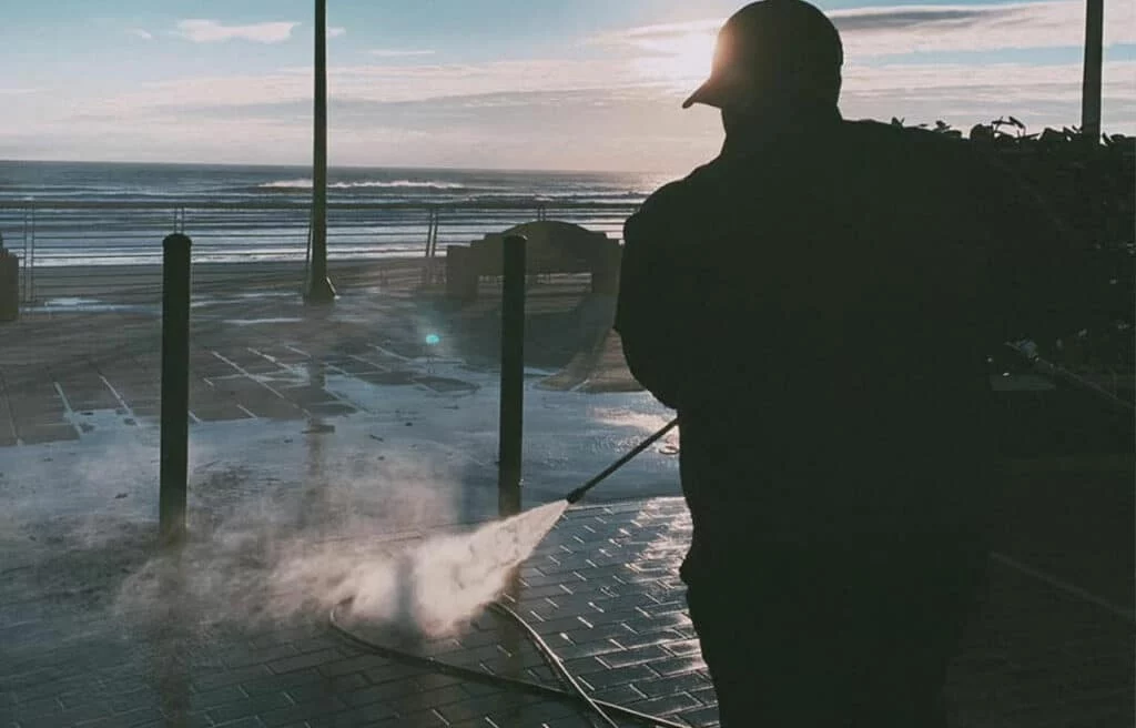 A worker power washes a brick promenade by the sea at sunset. Essential concrete cleaning removes grime, revealing clean surfaces and improving the coastal walkway.