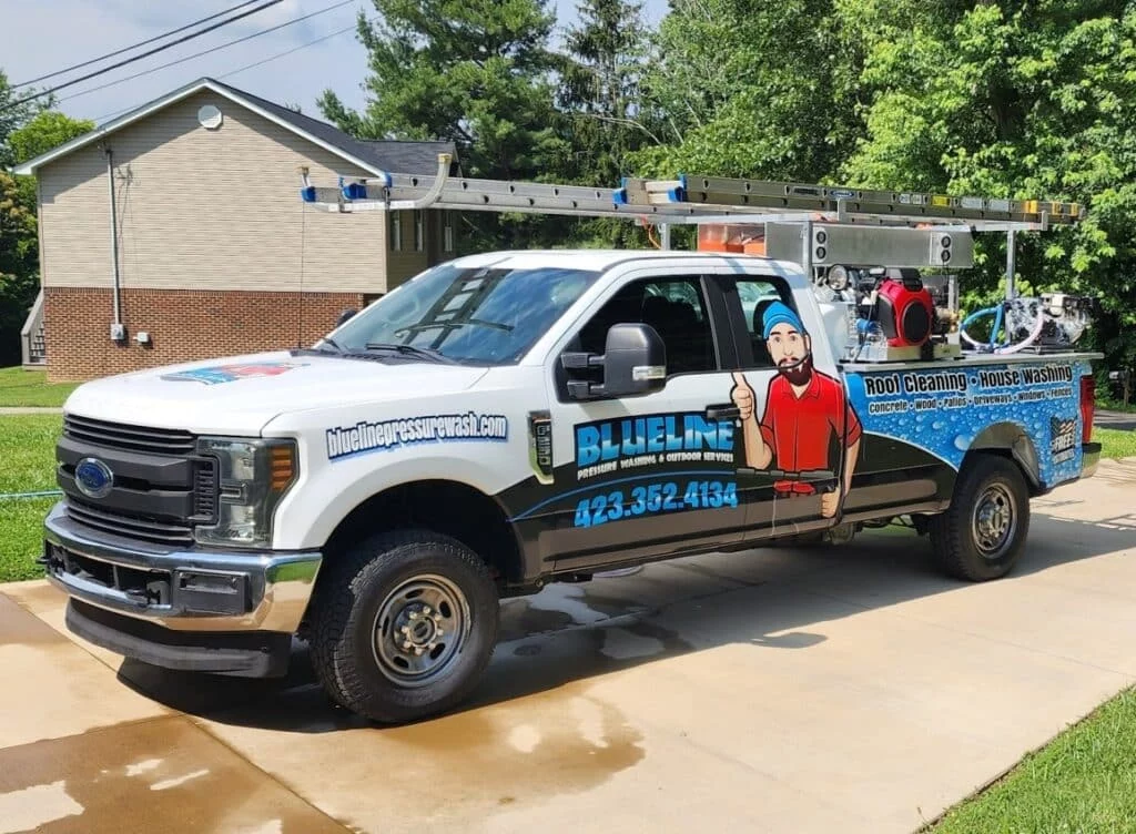A white truck, branded "Blueline Pressure Wash," is equipped for roof cleaning and house washing. The truck features a cartoon graphic and contact information. Ladder on top, ready for outdoor services.