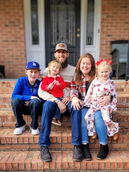 A family of five sits on brick steps in front of their house. The parents sit in the middle, with their three children beside and on their laps. The children are wearing festive holiday-themed clothing.