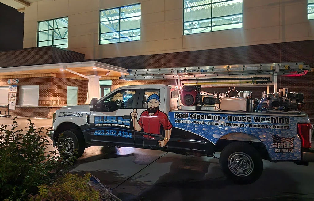 A Blue Line Pressure Washing truck with roof and house cleaning equipment parked in front of a brick building. Text on the truck includes "Roof Cleaning, House Washing, Concrete, Wood, Patios, Driveways, Windows, Fences" and the phone number 423-352-4134.