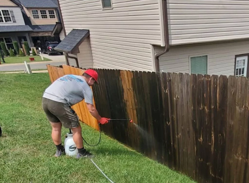 A man pressure washing a wooden fence in Johnson City TN. The fence is partially cleaned, showing the contrast between the dark, dirty wood and the bright, clean wood. End of summer pressure washing in action.