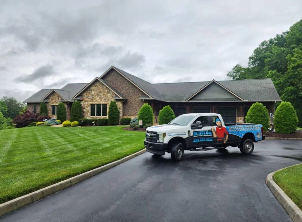 A pressure washing company truck parked on a driveway in front of a large, well-maintained home with a green lawn. The truck features the company logo and contact information. September is the best time for pressure washing.