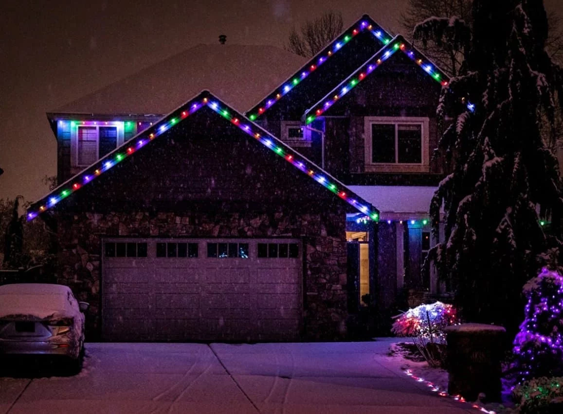 House decorated with colorful Christmas lights along the roofline in a snowy Tri-Cities neighborhood. A dusting of snow covers the driveway and car. Winter exterior cleaning is important to maintain homes.