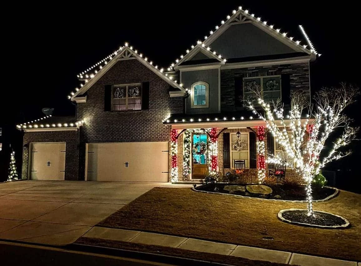 House with professional Christmas lighting at night. White lights line the roof and a tree in the yard, with red and white garlands on the porch columns.
