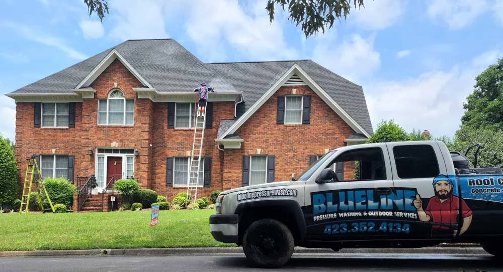 Blueline Pressure Washing truck parked in front of a large brick house. A worker on a ladder cleans the roof. Blueline offers pressure washing and deck staining services in Johnson City, TN. Call 423-352-4134.