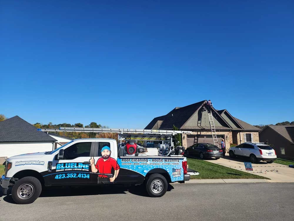 Blueline pressure washing truck parked on a suburban street in Johnson City, TN. A worker is on a ladder cleaning the roof of a house. "Blueline: Pressure Washing & Restoration, 423.352.4134. Roof Cleaning - House Washing.
