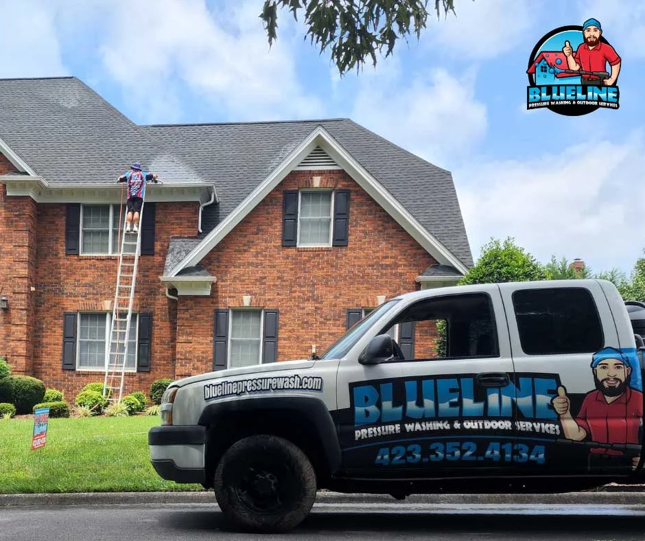 A worker on a ladder cleans gutters on a brick house. A truck for Blueline Pressure Washing & Outdoor Services is parked in front. Call 423-352-4134 for power washing services in Johnson City, TN.