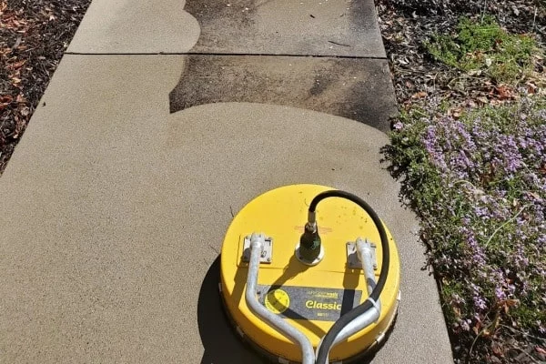 Power washing a concrete walkway in Piney Flats, TN. A yellow surface cleaner attached to a pressure washer removes dirt and grime, revealing a clean surface.