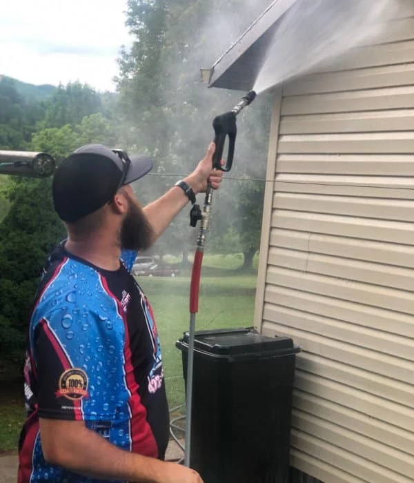 Man power washing a home's siding with a pressure washer and long wand. He wears a blue shirt with water droplet design and a cap. Black trash can nearby. Trees and mountains in the background.