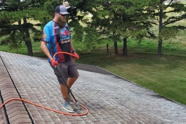 Man cleaning roof shingles with a power washer. He wears a branded shirt, shorts, and a cap. Trees and a house are visible in the background.