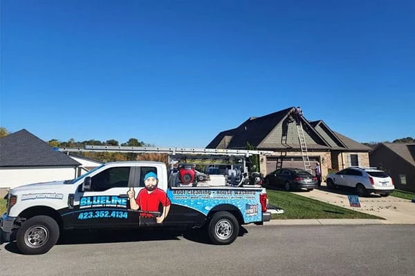 BlueLine roof cleaning truck parked in front of a home with a worker on the roof. Ladder is set up. Text on truck: "BlueLine Roof Cleaning - House Washing, 423.352.4134".