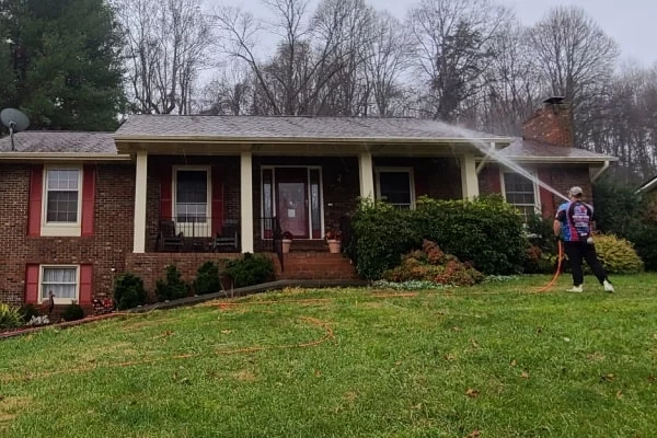 A man power washes the exterior of a red brick home with white trim, showcasing home maintenance. A green lawn and trees form the backdrop.