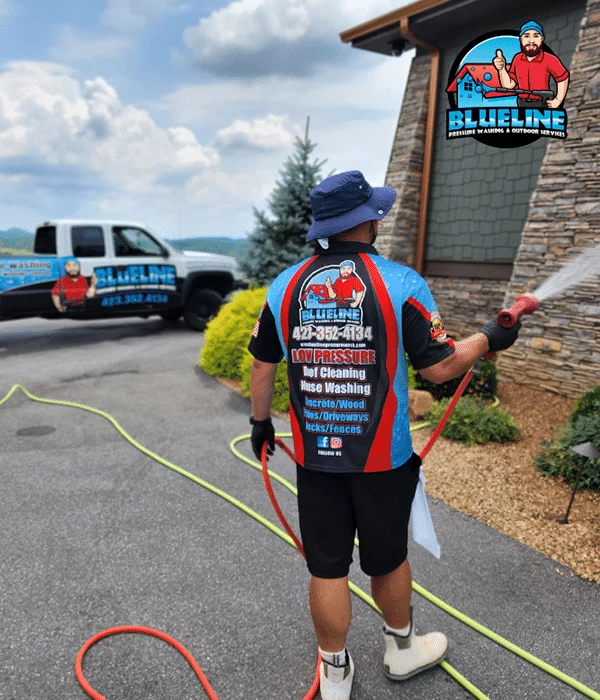 A pressure washing professional uses low pressure techniques to clean a stone house. The BlueLine Pressure Washing truck is visible in the background. Services include roof cleaning, house washing, and deck/fence cleaning.