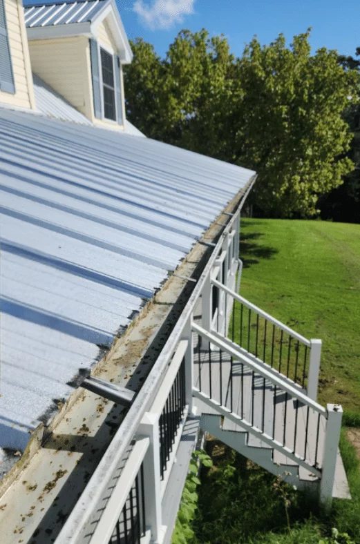 Gutter filled with leaves and debris running along the edge of a metal roof, highlighting the need for gutter cleaning and maintenance. A white staircase with black railings leads down from the gutter. Green lawn and trees in the background.