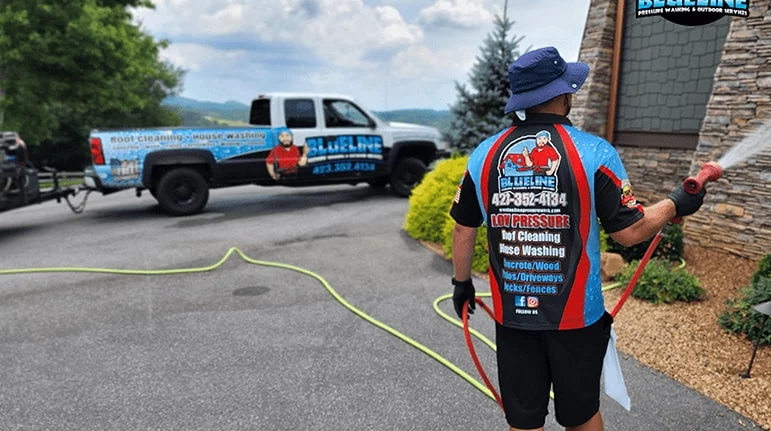 A Blueline Pressure Washing worker power washes a stone house. The company truck, advertising "Roof cleaning House washing 473.352.4134" is parked nearby. Low pressure, soft cleaning.