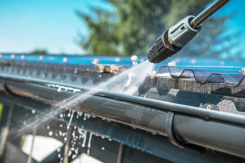 Close-up of a pressure washer cleaning dark gray gutters, water spraying forcefully to remove debris. A section of translucent corrugated roofing is visible above the gutter.
