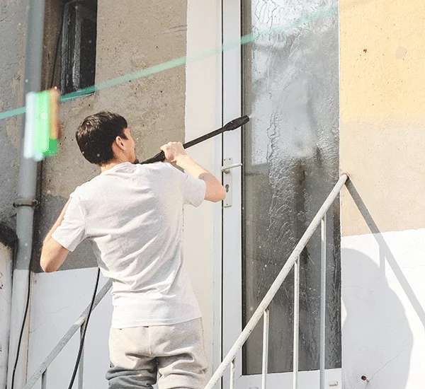A man power washing a white framed glass door on a light yellow building. He's wearing a white t-shirt and grey sweatpants, standing on metal stairs. The scene highlights the effectiveness of power washing.