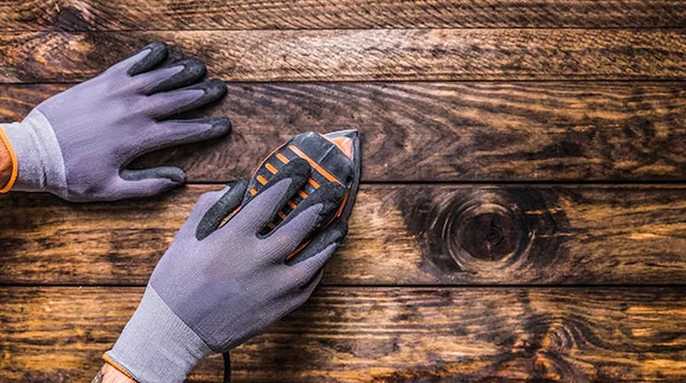 Close-up of gloved hands sanding a weathered wooden surface with a power sander. The wood grain is prominent, showcasing a rustic texture.