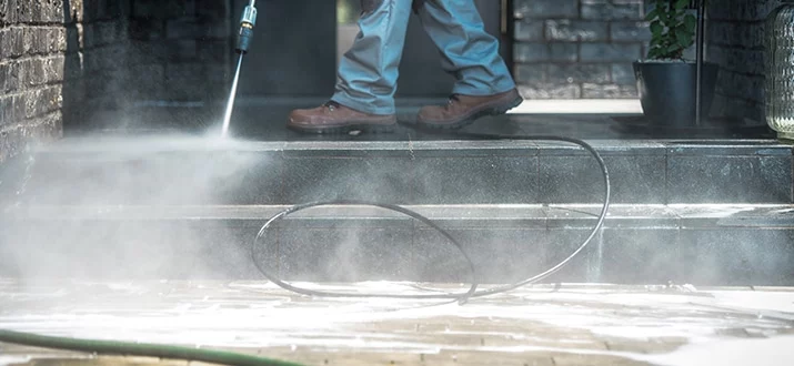 A person power washing the steps of a house, demonstrating how often a house should be power washed. The high-pressure spray blasts away grime, revealing clean stone beneath.