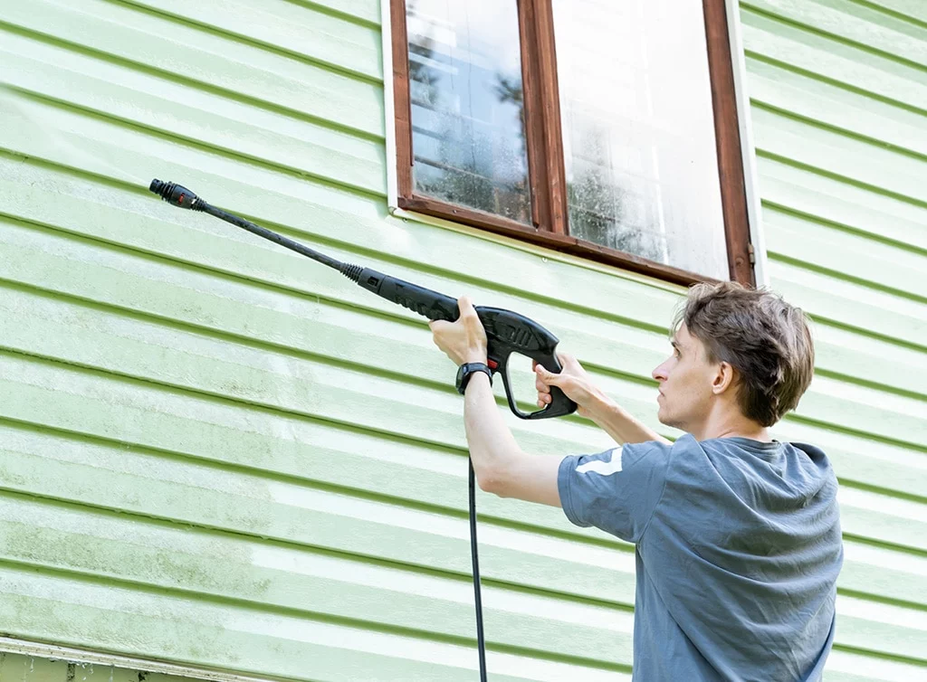 Man pressure washing green vinyl siding near a window. Cleaning exterior of a house to prevent damage.