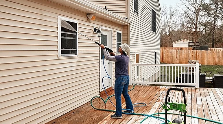 A woman pressure washing the siding of a house, standing on a wooden deck. She wears a hat and blue jeans, using a green pressure washer. Kingsport's #1 choice for pressure washing services.