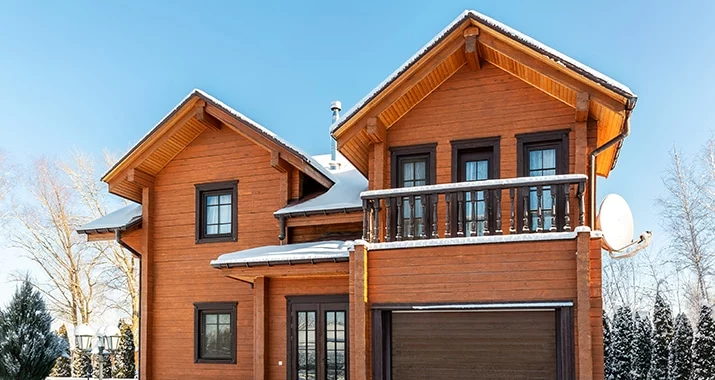 A two-story log cabin home with snow on the roof, set against a bright blue winter sky. A balcony with dark railings sits above the garage. Pressure washing would be ideal in warmer weather.