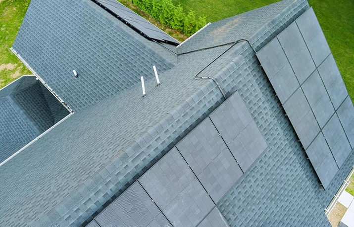 Aerial view of a house roof with dark gray asphalt shingles and solar panels. The panels are neatly arranged, capturing sunlight for energy efficiency.