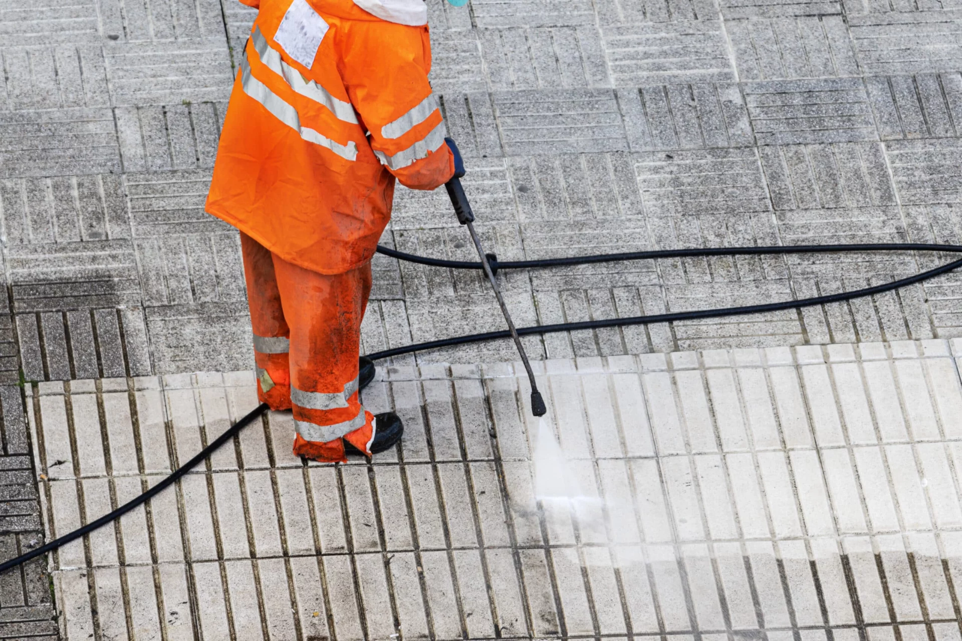 A worker in bright orange protective gear uses a pressure washer to clean a tiled sidewalk. The powerful spray reveals the clean tiles beneath, illustrating the effectiveness of pressure washing.