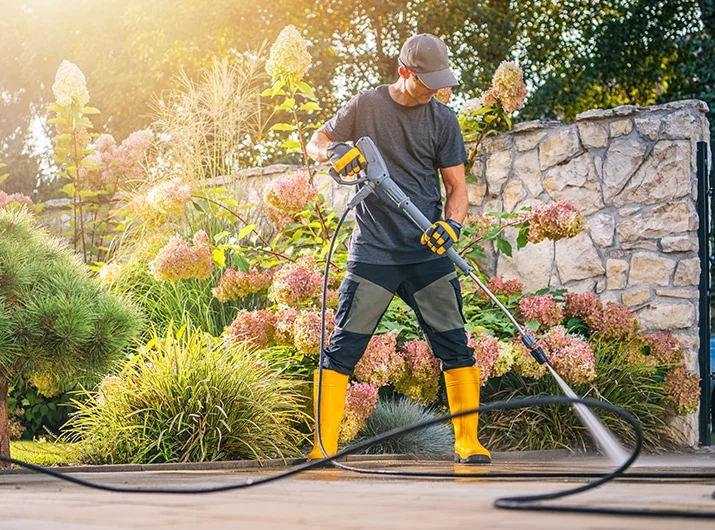 Man power washing a stone patio surrounded by hydrangeas. He wears yellow boots and uses a pressure washer to clean the surface, illustrating home maintenance.