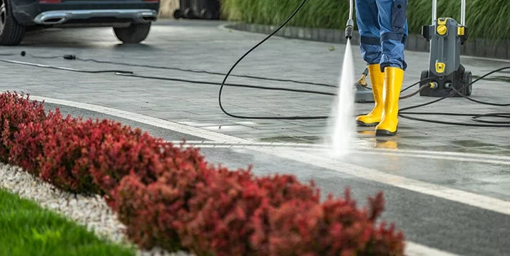 Person pressure washing a driveway with yellow boots. Red bushes line the edge, and a car is parked nearby. Pressure washing is an effective way to clean your home's exterior.