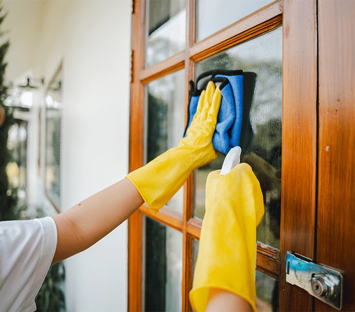 Person wearing yellow gloves cleans a window with a blue cloth and spray bottle. The wooden window frame is in focus, suggesting a detail shot of pressure washing preparation.