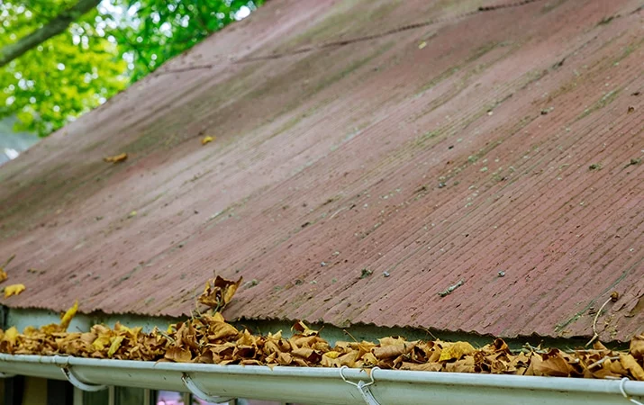 Clogged gutters overflowing with autumn leaves beneath a rusty, moss-covered roof. Pressure washing is essential for home maintenance, especially after fall.