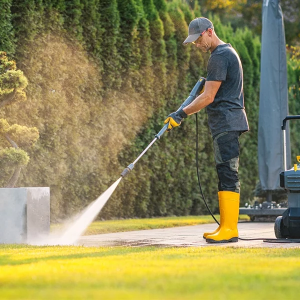 Man power washing a patio with a pressure washer, wearing yellow boots and gloves. The spray cleans a concrete block, showcasing the benefits of power washing outdoor surfaces. Green lawn and hedges in the background.
