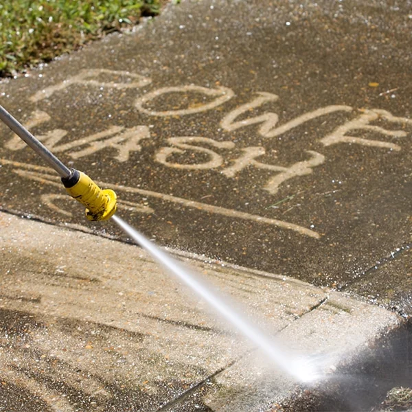 Power washing a concrete sidewalk with the words "POWER WASH" partially cleaned into the surface. A yellow pressure washer wand is pointed down at the concrete, spraying water.