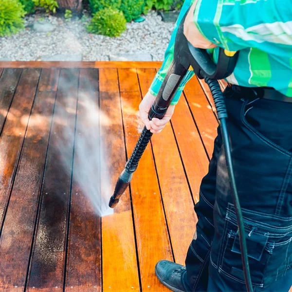 Close-up of a man power washing a wooden deck. The high-pressure water stream cleans the dark, weathered wood, revealing the fresh, lighter wood underneath. A demonstration of power washing techniques.