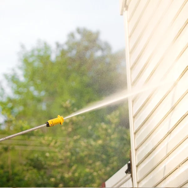 Power washing vinyl siding with a yellow pressure washer wand. Water sprays powerfully, cleaning the home's exterior. Trees are visible in the background.