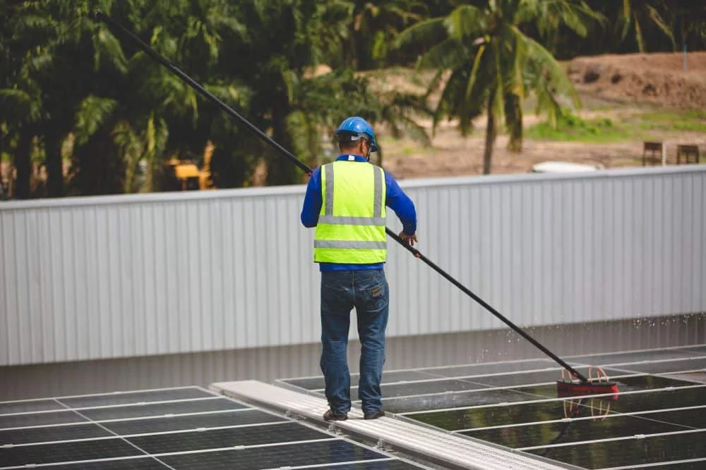 A worker in a hard hat and safety vest uses a long-handled brush for roof cleaning on solar panels atop a building. Palm trees are visible in the background.