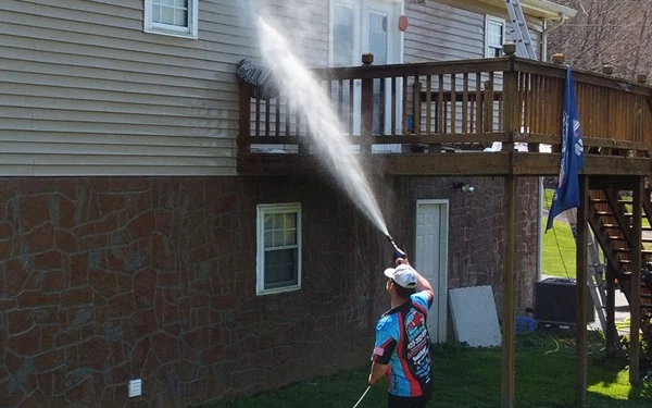 A man power washing a house exterior before repainting. The house has siding and a stone foundation, and he is spraying the siding with a high-pressure stream of water.
