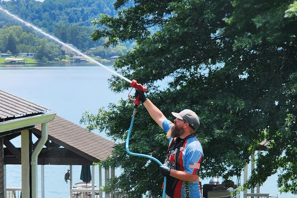 Man soft washing a house near a lake. He's using a long-range nozzle, demonstrating fence cleaning techniques. The mountain backdrop and lush greenery add to the scenic outdoor setting.