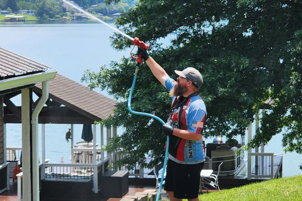 A man performs a Soft Wash roof cleaning, spraying a house roof with a long nozzle. The house is near a lake, with trees in the background. He wears a cap, gloves, and a patriotic-themed shirt.