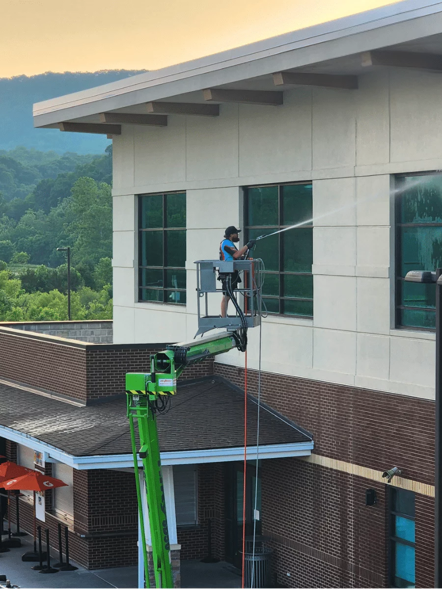 A worker power washing commercial building windows from a boom lift. The building has brick and light beige siding, with green trees visible in the background.