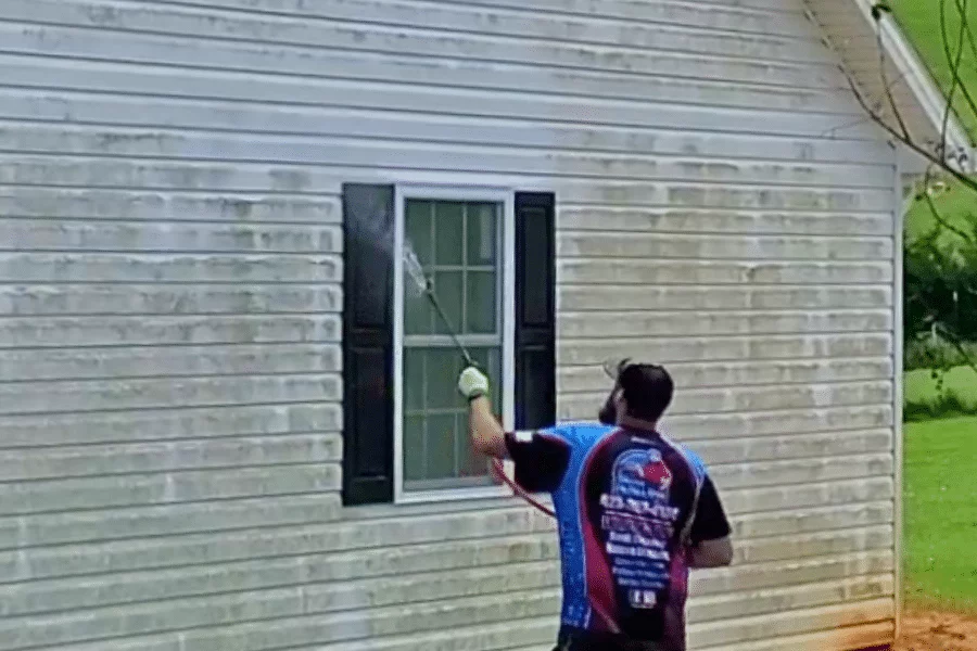A man soft washes a dirty, light-colored house with black shutters, using a long wand to spray cleaner on the siding.
