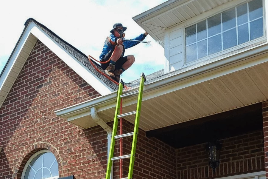 A worker soft washing gutters, perched on a roof with a ladder nearby. He's wearing safety gear and using a sprayer to clean the exterior of the home's gutters.