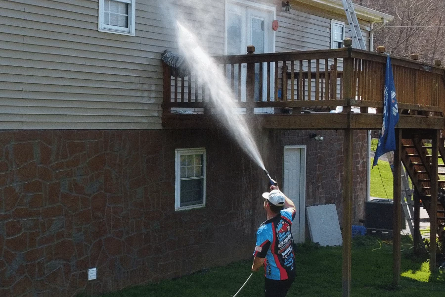 A man soft washing the side of a two-story house with a pressure washer. The house has tan siding and a stone foundation. He's aiming the stream toward a second-story deck.